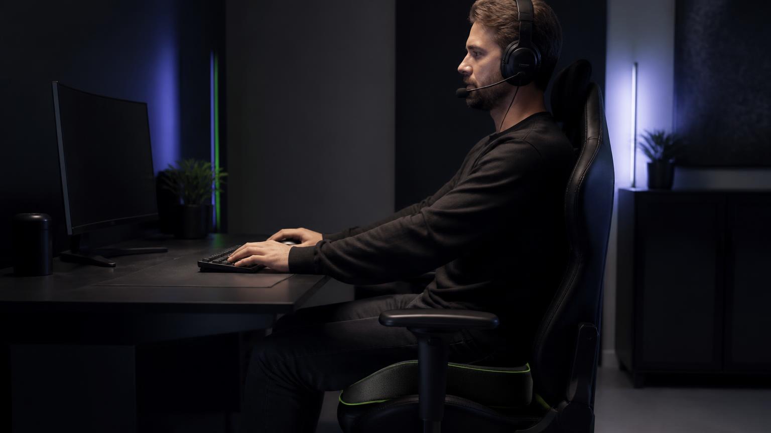 Gamer sitzt mit Headset auf einem schwarzen BackPal-Sitzkissen mit grüner Ziernaht an seinem Gaming-Setup